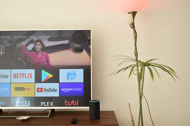Amazon Alexa acendendo uma lâmpada. Atrás de uma televisão mostrando a interface da Amazon Fire TV